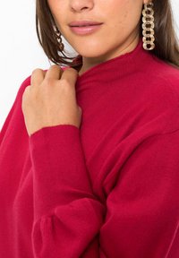 Maglione rosso rosa con collo alto e maniche lunghe, caratterizzato da una texture morbida e dettagli a coste sul colletto e sui polsini.