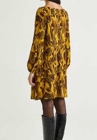 Robe à manches longues avec une base jaune ornée de motifs floraux marron. Design fluide avec une coupe décontractée, associée à des bottes noires montantes jusqu'aux genoux.