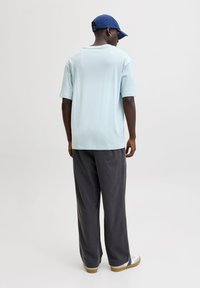 Lichtblauw shirt met korte mouwen en een ontspannen pasvorm, gecombineerd met donkergrijze, loszittende broek en witte sneakers met blauwe accenten, gezien van achteren.