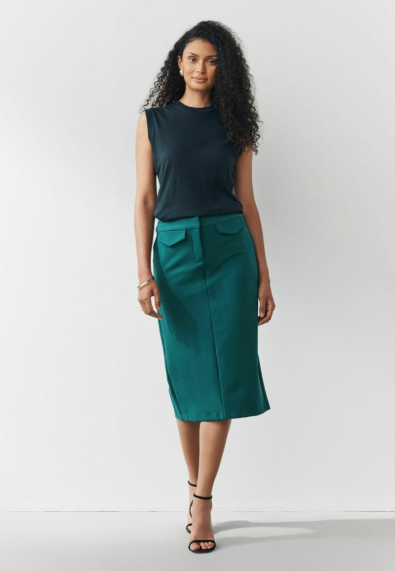 Top negro sin mangas combinado con una falda lápiz de teal a la altura de la rodilla que presenta bolsillos laterales y una textura suave. Sandalias negras de tiras.