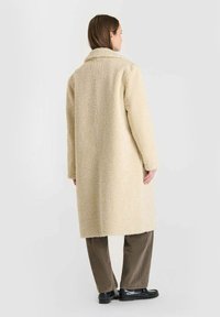 Beige teddyfrakke med en lang silhuet og stor krave, lavet af struktureret stof. Bæres over brune fløjl bukser og sorte sko.