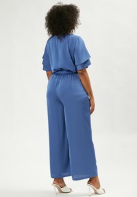Blauwe, zachte stof jumpsuit met fladdermouwen, elastische taille, wijd uitlopende broekspijpen en witte hoge hakken. Van achteren gezien.