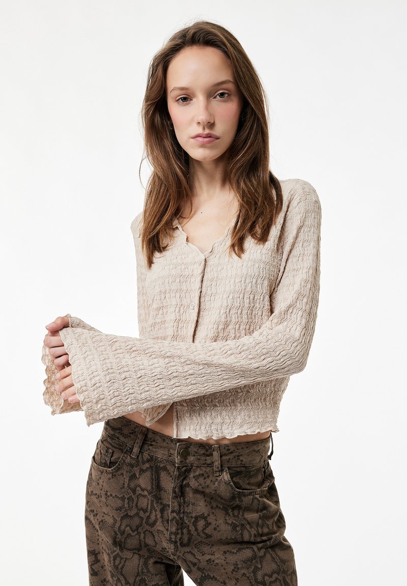 Cárdigan beige texturizado con mangas largas y acampanadas, con dobladillo festoneado y cierre con botones. Llevado sobre pantalones marrones con estampado de serpiente.