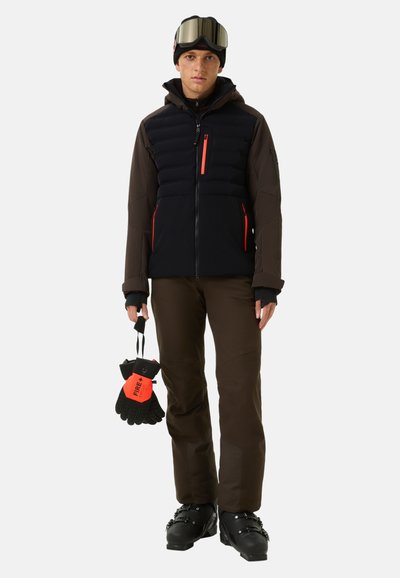 Ski jacket with black puffy front and brown sleeves, paired with brown pants. Holds black and orange gloves. Wears ski goggles and a beanie.