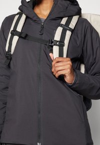 Czarna kurtka na zamek z teksturowanej tkaniny z kapturem, zestawiona z beżowym plecakiem w kratkę i regulowanymi paskami.