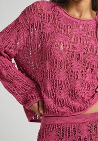 Różowy sweter z dzianiny o luźnym kroju, z teksturowanym wzorem z otwartych oczek oraz prążkowanymi mankietami i dołem, zapewniającymi przewiewny fason.
