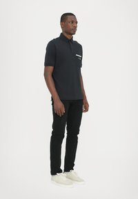 Svart polotrøye med brystlomme og hvit kant, parret med svarte slim-fit jeans og lysegrå sneakers. Enkel og moderne design.