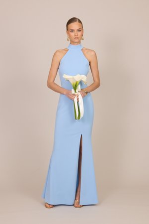Femme en robe bleu clair sans manches tenant des calla blancs avec un ruban, debout sur fond beige.