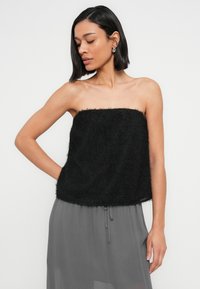 Γυναίκα με κοντά σκούρα μαλλιά, που φοράει ασημένια σκουλαρίκια κρίκους, μαύρο αμάνικο χνουδωτό τοπ και γκρι φούστα με κορδόνι, ποζάροντας με το ένα χέρι λυγισμένο.