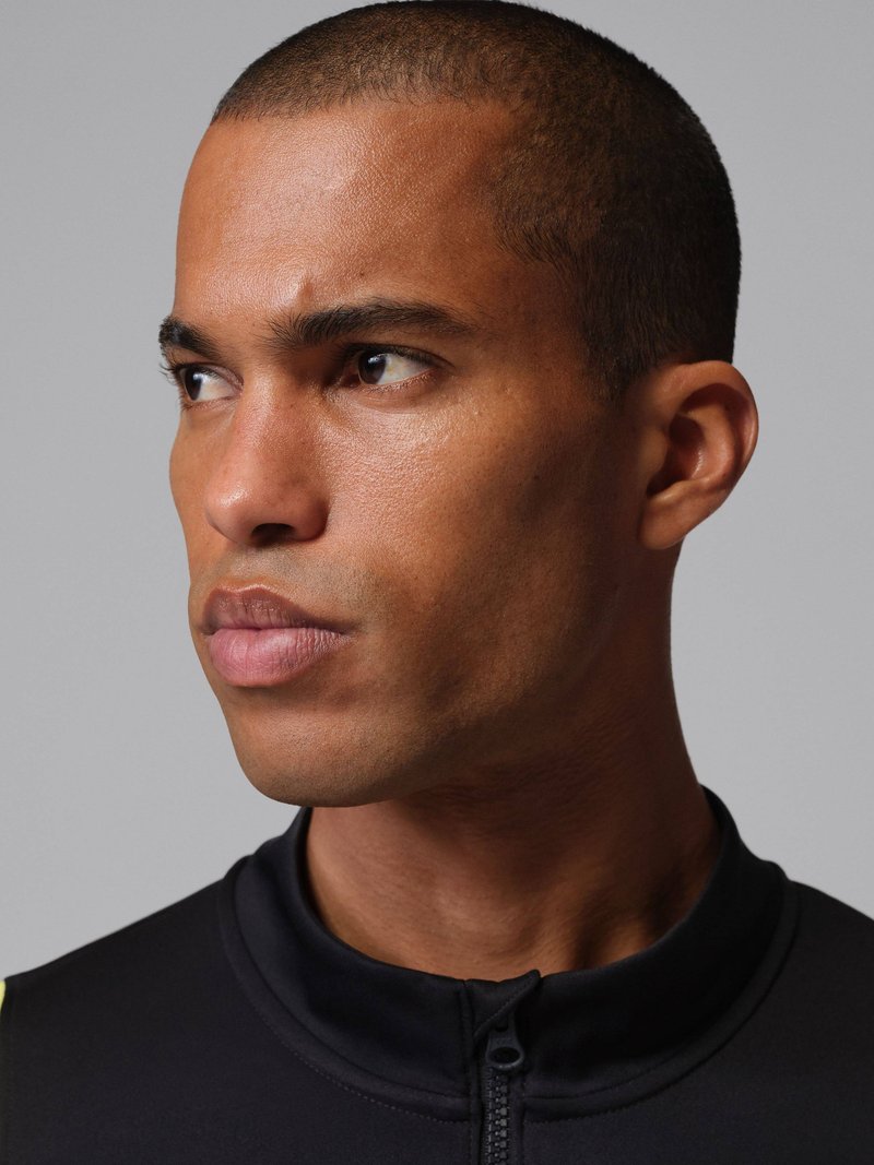 Hombre de cabello corto mirando hacia la izquierda, vestido con una prenda negra con cremallera, sobre un fondo gris liso.