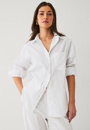 Chemise blanche en lin à boutons avec col, manches retroussées, poche poitrine unique et coupe légèrement décontractée. Texture lisse, design minimaliste.