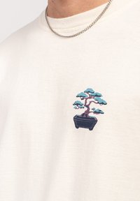 Mężczyzna w białej koszuli z małym nadrukiem drzewa bonsai na piersi oraz srebrnym naszyjnikiem w formie łańcuszka.