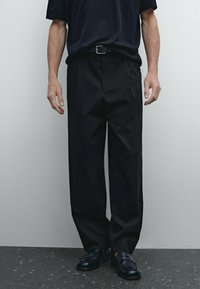 Black, pleated trousers worn with a dark shirt, featuring a belt. The fabric appears smooth, with a loose fit extending to the ankles.