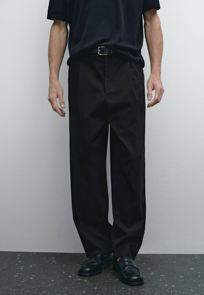Black, pleated trousers worn with a dark shirt, featuring a belt. The fabric appears smooth, with a loose fit extending to the ankles.