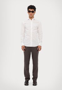 Witte button-up shirt, grijs gestreepte broek en zwarte schoenen. Model draagt donkere zonnebril. Schone, op maat gemaakte pasvorm met lange mouwen.