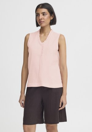 Kvinde iført en ærmeløs lyserød bluse med flæset V-hals og mørke, knælange shorts, stående mod en ensfarvet baggrund.