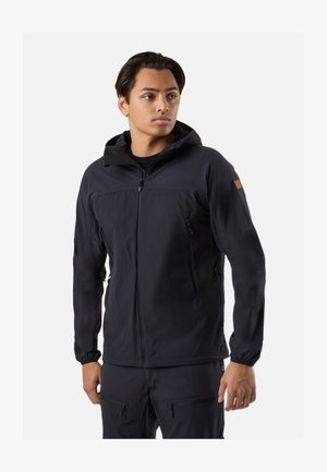 Jonge man die een zwarte hooded jas met rits en ritssluitingen op de zakken draagt, kijkt iets zijwaarts tegen een effen witte achtergrond.