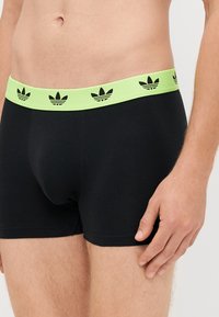 Mustat bokserit limevihreällä joustavalla vyötäröllä, jossa on toistuva musta Adidas-logokuvio. Sileä kangas ja istuva muoto.
