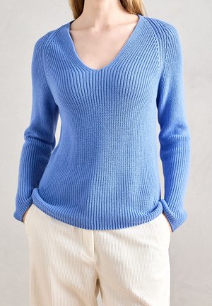 Frau trägt einen blauen, gerippten V-Ausschnitt-Pullover mit langen Ärmeln und cremefarbene Hose, Hände in den Taschen, neutraler Hintergrund.