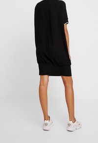 Černé pletené šaty s volným střihem, krátkými rukávy a žebrovaným lemem. Nošeny s bílými teniskami, ukazující hladkou texturu látky.