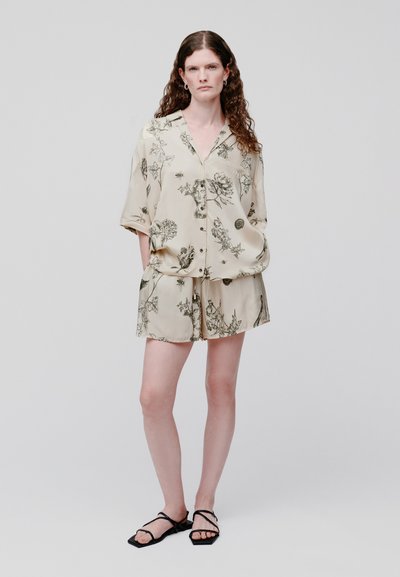 Femme aux longs cheveux bouclés portant une chemise beige boutonnée et un short assorti avec des imprimés botaniques noirs, debout les mains dans les poches.
