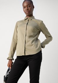 Camisa de mangas longas verde-oliva, com colarinho, com placket de botões pretos e punhos franzidos; combinada com calças pretas e segurando uma mala preta.