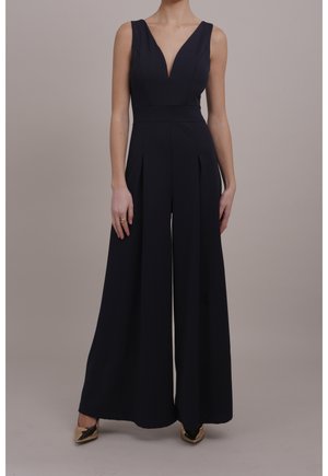 Vrouw die een zwarte mouwloze jumpsuit draagt met een diepe V-hals en wijde pijpen, gecombineerd met gouden puntige hakken.