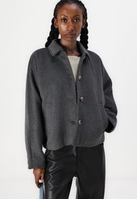Veste en mélange de laine gris avec un col, cinq boutons, coupe décontractée et texture subtile. Associée à un pantalon en cuir noir.