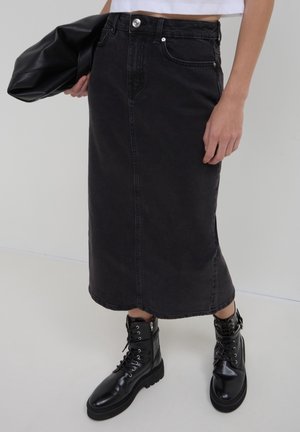 Frau trägt einen schwarzen, hoch taillierten midi Jeansrock, schwarze Schnür-Combat-Stiefel und hält eine schwarze Leder-Clutch.