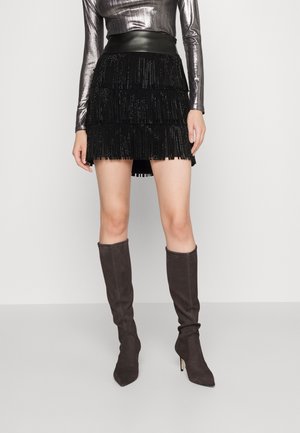 Jupe mini en franges noires avec une texture superposée, associée à des bottes en daim grises, montantes jusqu'au genou, avec des bouts pointus et des talons aiguilles.