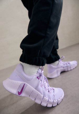 Personne portant des chaussettes blanches, un pantalon noir et des baskets Nike violet clair avec des semelles segmentées, marchant sur une surface de sol neutre.