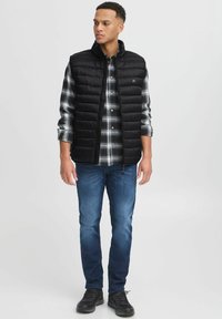 Gilet piumino nero con cuciture orizzontali su una camicia di flanella a scacchi grigi e neri, abbinato a jeans blu e sneaker nere.