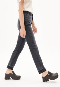 Jean en denim noir à coupe slim, ourlet légèrement effiloché, associé à des sabots noirs à enfiler avec un talon chunky et un motif ondulé.