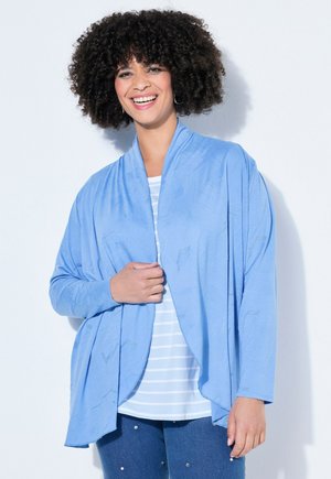 Smilende kvinde med krøllet hår iført en lys blå åben cardigan over en hvid stribet skjorte og blå jeans med små dekorationer.