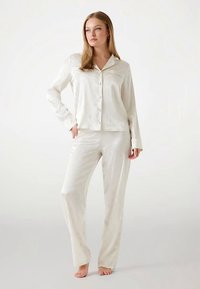 Ensemble de pyjama en satin couleur crème comprenant une chemise boutonnée avec une poche et un pantalon long, tous deux à la texture lisse et à la coupe ajustée.