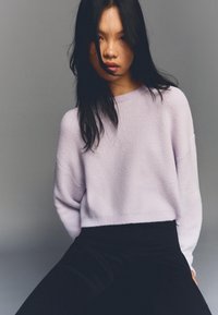 Svetlo vijoličen teksturiran pulover z okrolim izrezom in spuščenimi ramenih, kombiniran s črnimi hlačami z visokim pasom, postavljen proti nevtralnemu ozadju.