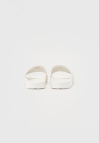 Des sandales en caoutchouc blanches à semelle plate, avec de larges lanières et une texture lisse. La semelle est légèrement surélevée avec un motif de grip texturé.