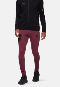 Mann in burgunderfarbenen Sportleggings mit schwarzem seitlichem Taschen und schwarzen Wanderschuhen, kombiniert mit einer schwarzen Reißverschlussjacke.
