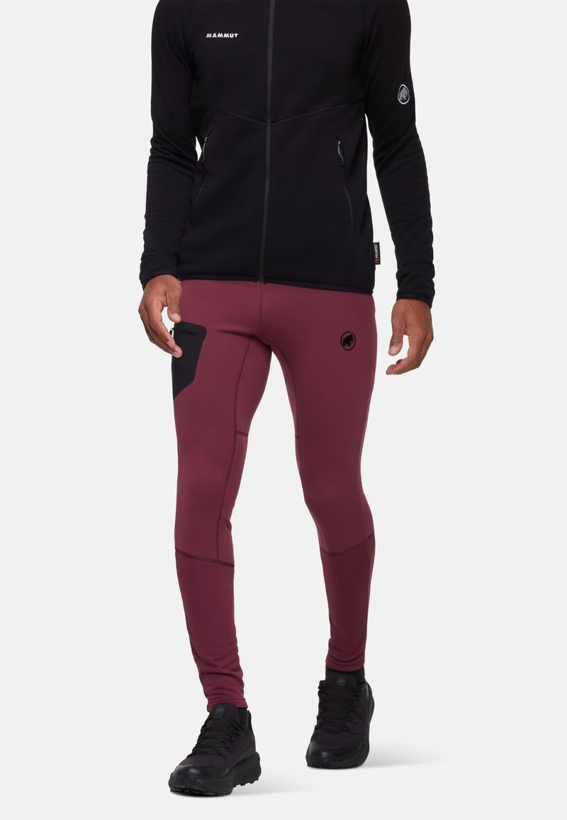Mann in burgunderfarbenen Sportleggings mit schwarzem seitlichem Taschen und schwarzen Wanderschuhen, kombiniert mit einer schwarzen Reißverschlussjacke.