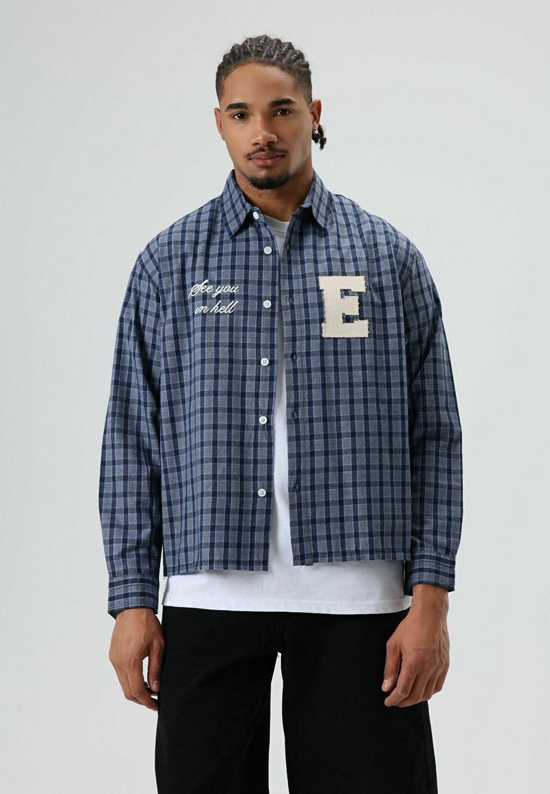Blauw en grijs geruit overhemd met knoopsluiting, borstlap met 'E' en geborduurde tekst "see you in hell," gedragen over een wit T-shirt.