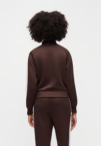 Καφέ φόρμα με ψηλό γιακά, με μπεζ χρωματικές εναλλαγές στα μανίκια. Λείο ύφασμα και εφαρμοστό σχέδιο.