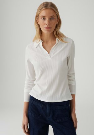 Blond meisje met een wit geribd poloshirt met lange mouwen en donkerblauwe spijkerbroek, staand tegen een effen lichte achtergrond.