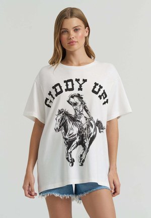 Young woman wearing oversized white T-shirt with black cowboy riding horse graphic and "GIDDY UP!" text, paired with denim shorts.