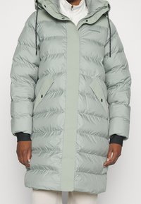 Ljusgrön puffjacka med huva, vertikal söm och två framfickor. Har ribbade ärmslut och en slät, vadderad textur.