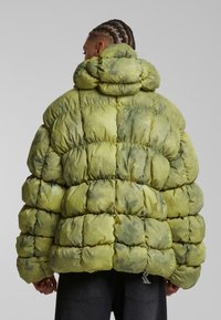 Overdimensioneret pufferjakke i lys grøn med tie-dye effekt, der har quiltede sektioner og høj krave. Snøre i kanten.