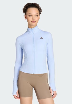 Femme aux cheveux bouclés portant une veste de sport bleu clair zippée et un short de cyclisme beige ajusté, debout avec les mains détendues le long du corps.