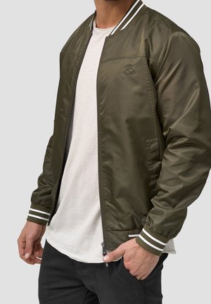 Mand iført olivengrøn bomberjakke med hvide stribede manchetter og krave, hvid t-shirt og sorte bukser mod en grå baggrund.