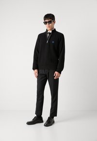 Pullover nero a maglia con colletto con zip ed emblema blu, abbinato a pantaloni neri su misura e scarpe di pelle nere. Occhiali da sole neri indossati.