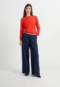Un pull rouge à col rond en matière douce, associé à un pantalon en denim noir à jambes larges. L'ensemble offre une coupe propre et ajustée.