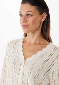 Pletený cardigan v krémové barvě, se scalloped výstřihem, vertikálními pruhy a knoflíkovým zapínáním s malými dírkami pro ventilaci.
